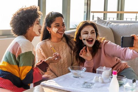 Multicultural friends applying facial masks and taking selfie on cozy skincare night at home