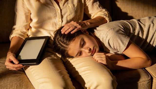 Mother comforting teenage daughter on sofa with e-reader during quiet evening at home