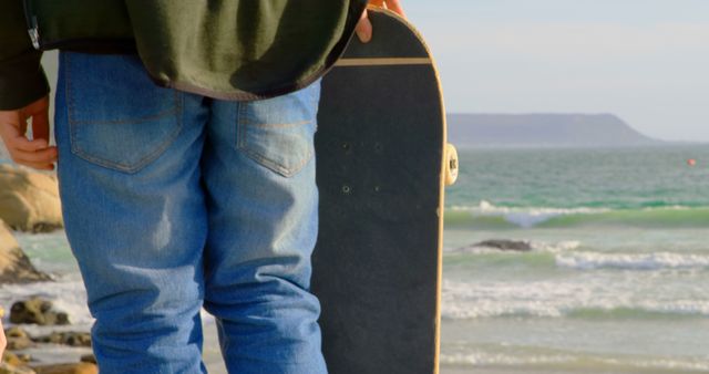 Person wearing jeans holding skateboard while standing on beach, with ocean and horizon visible. Perfect for lifestyle blogs, outdoor adventure promotions, skateboarding community websites, and marketing materials for casual and sporty clothing brands.