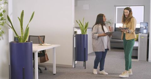 Diverse female coworkers discussing project in modern open-plan office holding tablets