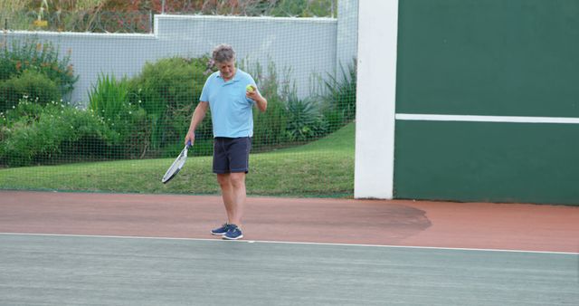 Senior man holding tennis ball while preparing for a serve on tennis court. Ideal for ads related to sports, senior fitness, tennis equipment promotions, and active lifestyle campaigns aimed at older adults.