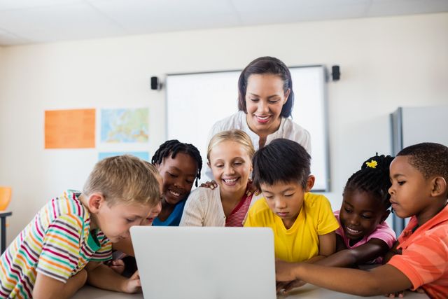 This image shows a diverse group of children gathered around a laptop with their teacher in a classroom. They are engaged and happy, indicating a positive learning environment. This image can be used for educational websites, school brochures, online learning platforms, and articles about modern teaching methods and technology in education.