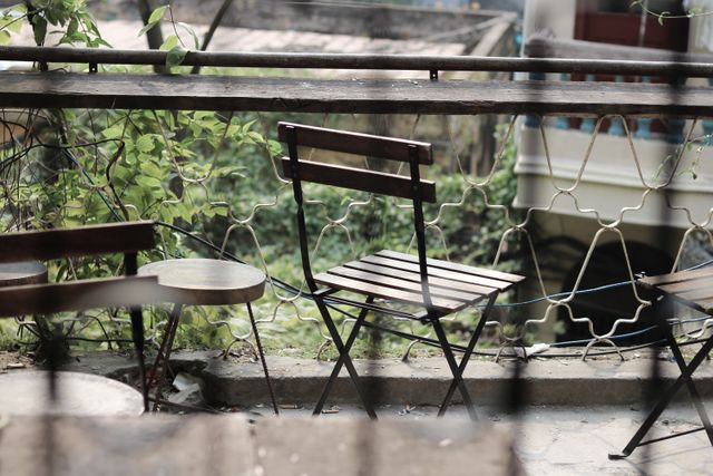 Empty outdoor cafe chairs and tables on a rustic balcony offer a nostalgic and peaceful atmosphere. The urban setting with greenery in the background is ideal for portraying leisurely moments, philosophical reflection, or promoting cafe culture. The scene can be used to suggest solitude, tranquility, or an invitation to pause and relax in marketing materials or editorial content.