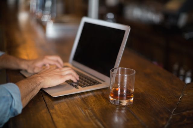 Man typing on laptop at pub with drink nearby. Ideal for illustrating remote work, casual business settings, or leisure activities. Useful for articles on work-life balance, technology in everyday life, or the modern work environment.