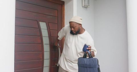 Man reaching for front door while talking on smartphone carrying bag and travel mug