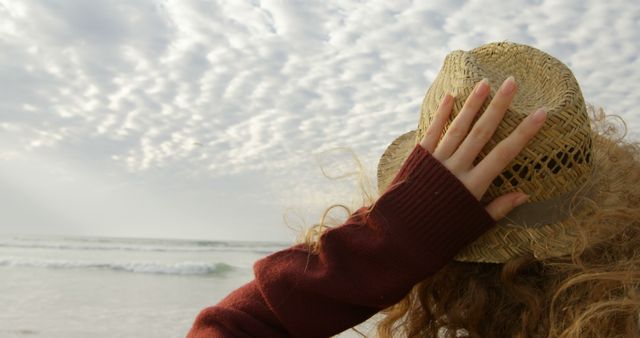 A young woman stands at the beach, holding her straw hat while looking towards the ocean and cloudy sky. Ideal for promoting travel destinations, relaxation products, vacation packages, peaceful moments, nature retreat brochures or coastal tourism industries.