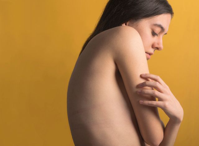 Young woman with bare shoulders standing against yellow wall, holding herself gently. Useful for mental health, serenity, beauty, self-care, and contemplative or reflective themed content.