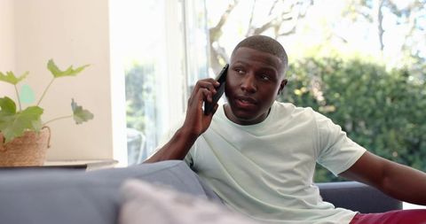 Talking on phone while relaxing on sofa near sliding door with sunlight and houseplant
