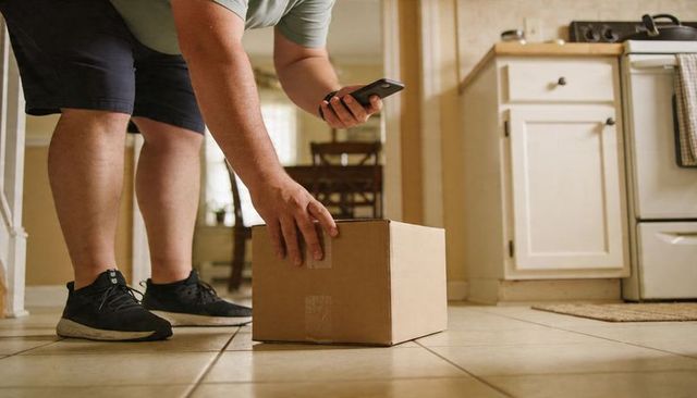 Bending person scanning parcel with smartphone on kitchen floor during home delivery