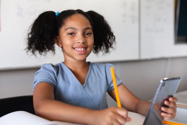 Young biracial schoolgirl smiling while using a digital tablet at her desk in a classroom. Ideal for educational materials, technology in education promotions, and school-related content. Perfect for illustrating modern learning environments and student engagement with digital tools.