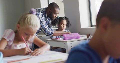 Male Teacher Leaning Over Desk Guiding Young Student with Workbook in Bright Elementary Classroom