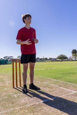 Teen cricketer standing on pitch holding ball near stumps on sunny training ground
