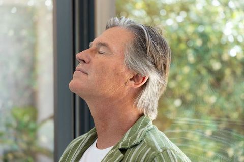 Mature man standing with eyes closed by sunlit window contemplating calm mindful moment