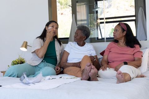 African american senior women applying skincare on bed sharing self-care routine and bonding
