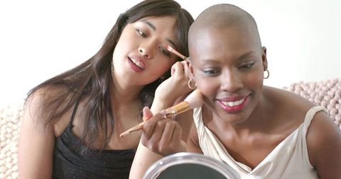 Multicultural women applying makeup on sofa using tabletop mirror and brushes for beauty