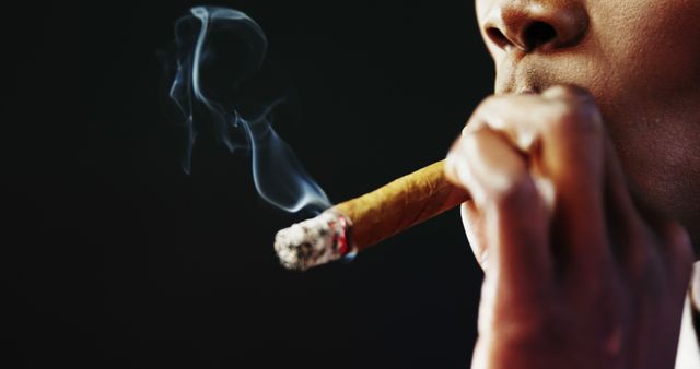 Close-up of a man smoking a cigar with a thin stream of smoke trailing. Suitable for use in lifestyle articles, luxury brand promotions, health awareness campaigns discussing smoking habits, or illustrating indulgence and relaxation activities.