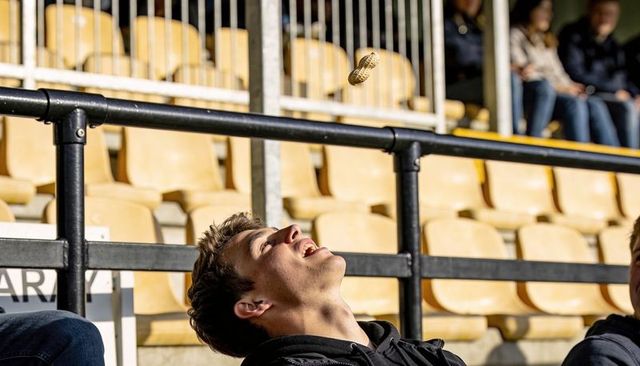 Leaning young man catching peanut midair in sunlit stadium bleachers