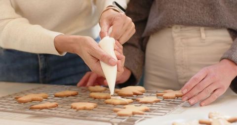Female friends decorating homemade flower-shaped cookies with piping bag on cooling rack
