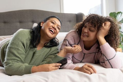 Indian and african american friends laughing lying on bed in cozy knit and grid sweaters