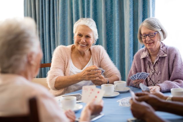 Senior women are gathered around a table, enjoying a card game and coffee in a nursing home. They are smiling and engaging in a friendly and relaxed atmosphere. This image can be used for promoting senior living communities, retirement homes, social activities for the elderly, and the importance of friendship and leisure in later life.