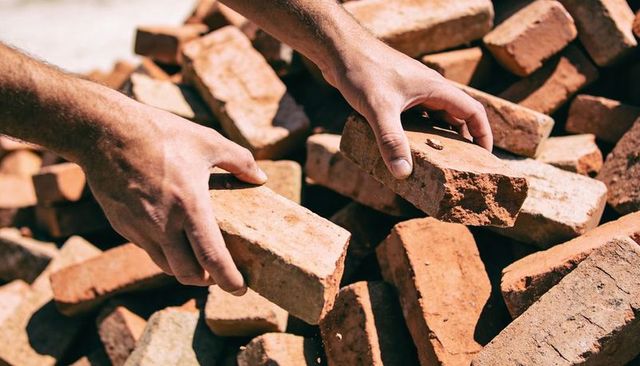 Worker lifting worn clay bricks with bare forearms from sunlit masonry pile