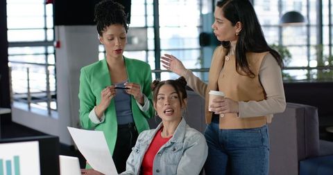 Multiracial women collaborating over documents and monitor in modern open-plan office