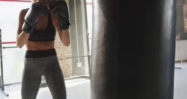 Athletic woman wearing grey leggings and black sports bra practicing boxing with punching bag. Ideal for fitness, exercise, and healthy lifestyle projects. Great for illustrating concepts of strength, training, and physical fitness. Can be used for gym promotions, sports equipment advertisements, and motivational health campaigns.