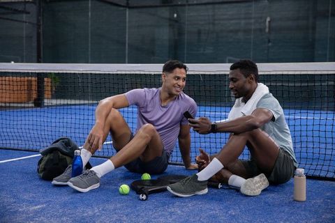 Diverse male friends resting on blue court sharing smartphone after paddle match