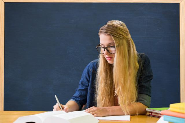 Young woman with glasses studying at desk with an open book, blackboard background. Ideal for promoting educational content, study tips, school advertisements, academic guides, or back-to-school campaigns.