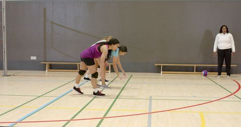 Female volleyball players practicing ready reception stance in indoor gym with coach