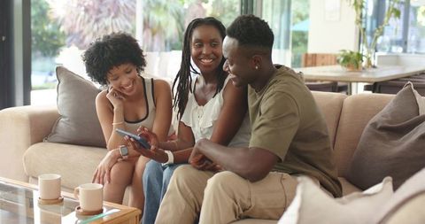 African American friends laughing and sharing smartphone while relaxing on sunlit sofa