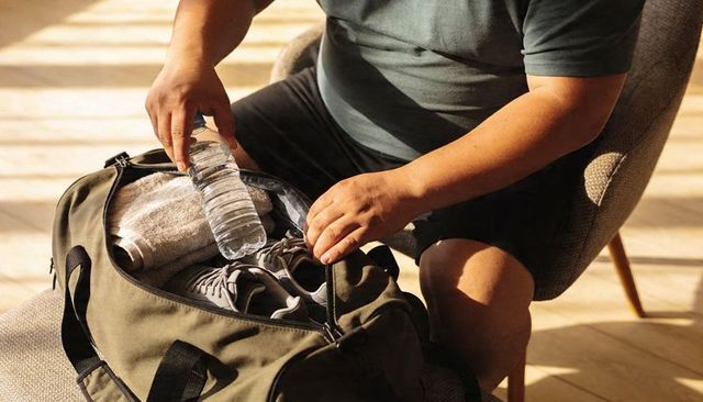 Man packing gym bag placing water bottle and sneakers into olive duffel in warm sunlight