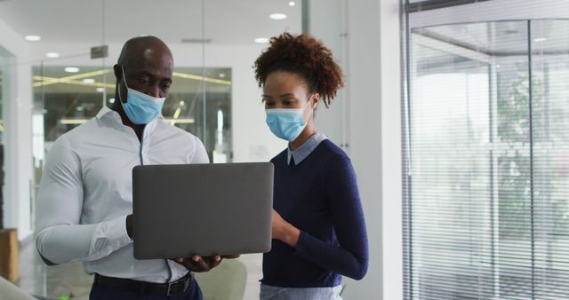 Two professionals wearing face masks collaborating on a project using a laptop in a modern office. Ideal for illustrating COVID-19 safety measures in a workplace, teamwork, and technology use. Useful for articles on office collaboration during pandemic, business environments, and health safety protocols at work.