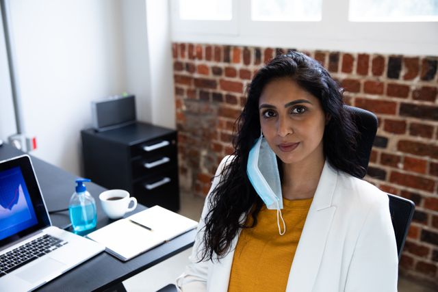 Portrait of biracial woman working in a casual office, looking at camera and smiling, wearing face mask. Social distancing in the workplace during Coronavirus Covid 19 pandemic.