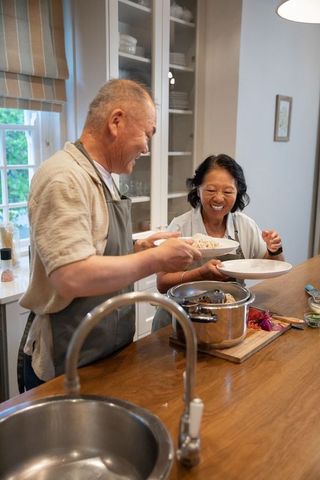 Asian senior couple preparing and serving homemade meal at sunlit kitchen island sharing joy