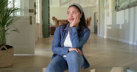 Pensive woman sitting at entryway resting elbow on knee in denim jacket and headband