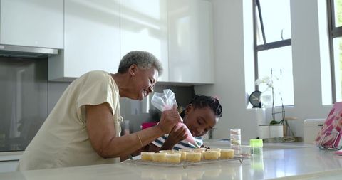Grandmother and granddaughter decorating cupcakes together on sunlit kitchen island