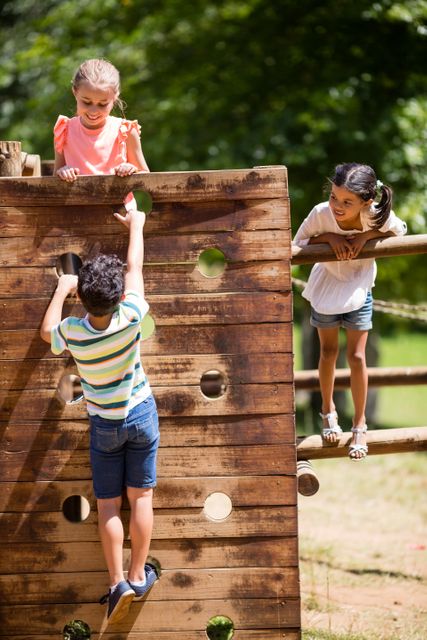 Children enjoying outdoor activities on a wooden playground structure in a park on a sunny day. Ideal for use in content related to outdoor play, childhood development, summer activities, and promoting healthy lifestyles for kids.