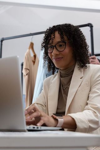 African american woman typing on laptop in boutique workspace surrounded by clothing