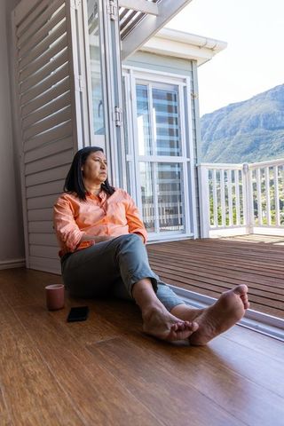 Asian woman sitting at sliding door to deck with mountain view, smartphone and mug