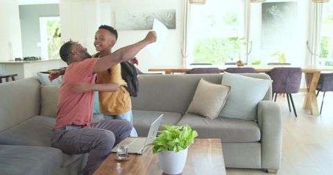 African american father hugging teenage son at home with backpack and laptop on table