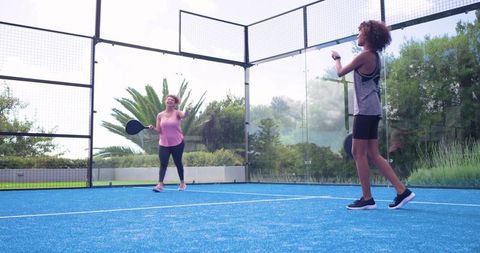 African american women playing padel on blue outdoor glass-enclosed court