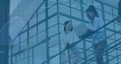 Businesswomen Collaborating on Tablet in Glass Staircase with Financial Charts Overlay