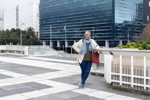 Senior african american woman leaning on railing with red tote in urban plaza, casual downtown