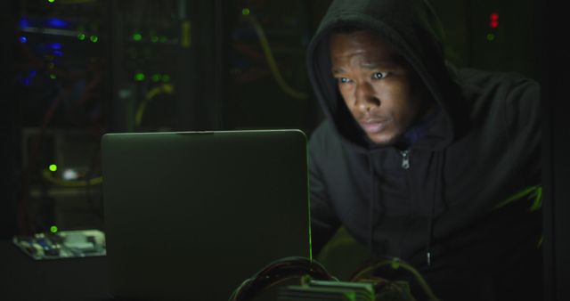 African American man deeply focused, working on laptop in dark server room. Suitable for content on cybersecurity, cybercrime, technology, coding, data security, and digital crime. Ideal for illustrating tech professionals monitoring security threats.