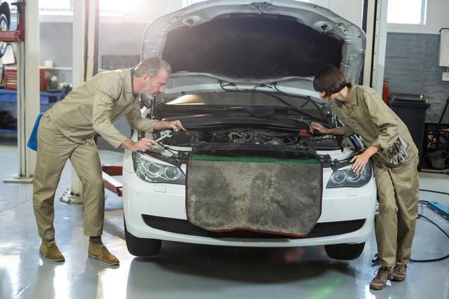 Mechanics wearing uniforms examining car engine in auto repair shop. Suitable for content related to auto repair services, vehicle maintenance, professional mechanics, and car workshops. Useful for illustrating teamwork, technical expertise, and routine car maintenance.