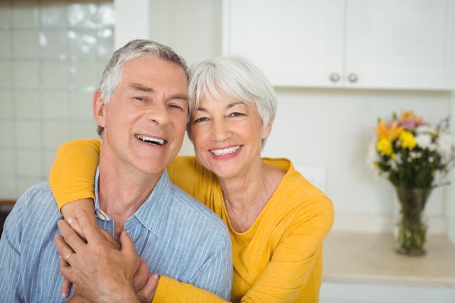 Senior couple embracing and smiling in a bright kitchen with flowers in the background. Perfect for use in advertisements, articles, and promotions related to senior living, retirement, relationships, and home life.