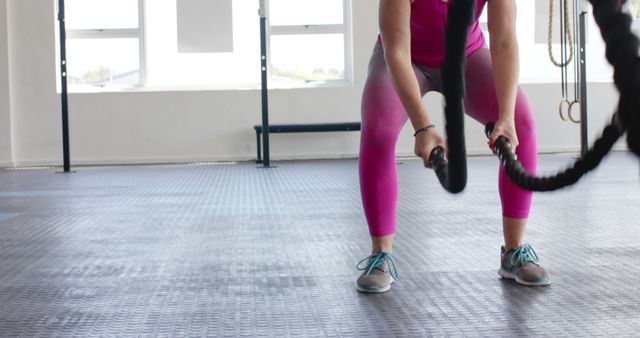 Fit woman exercising in a modern gym using battle ropes for strength training. Wearing vibrant pink leggings and sneakers, she demonstrates a powerful movement routine. Perfect for fitness blogs, workout guides, gym promotions, or ads focused on an active and healthy lifestyle.