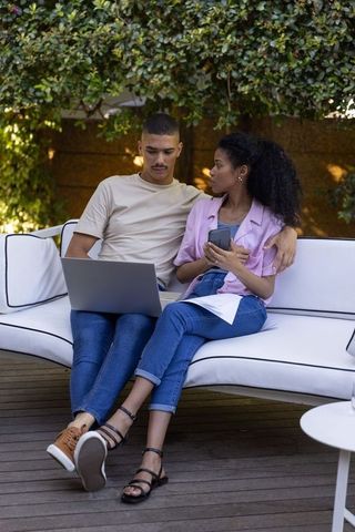 Diverse couple planning and working together on laptop and smartphone on outdoor deck sofa