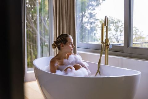 Woman relaxing in bubble bath by window with brass faucet in minimal modern bathroom